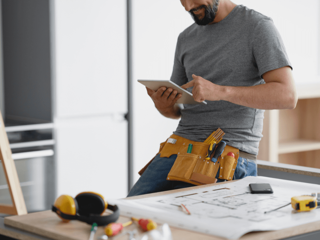 Tradesman using a digital tablet while seated at a table with project plans and tools.